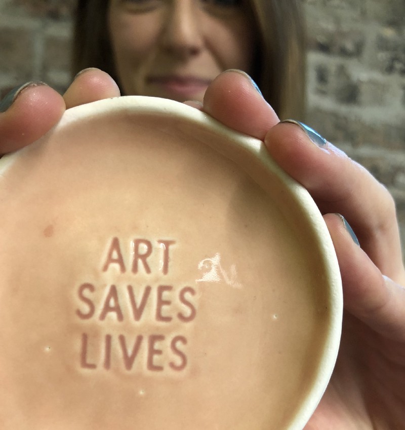 Rachel Hammond Holding a small ceramic plate reading "ART SAVES LIVES"
